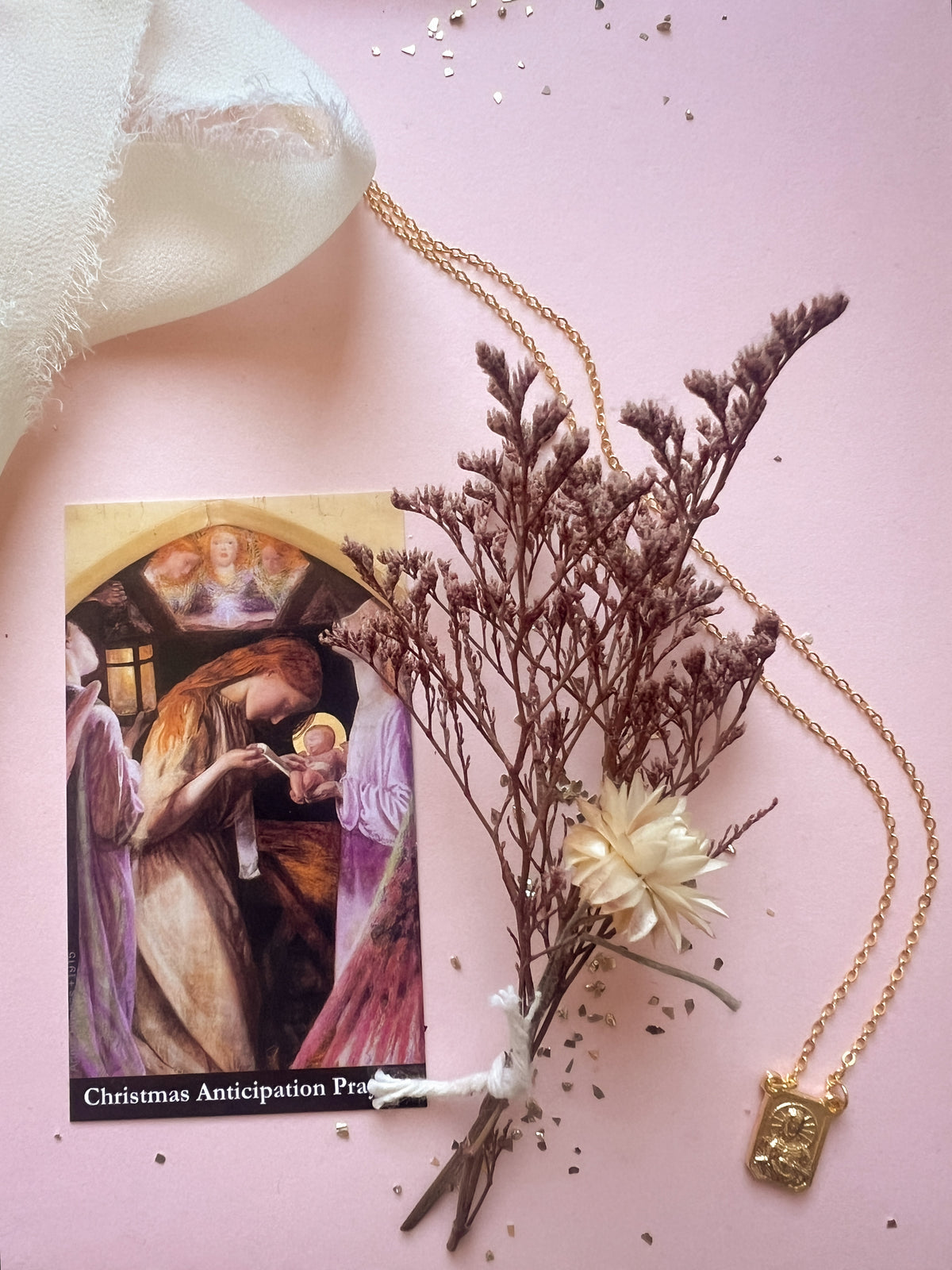 A gold-toned Scapular necklace with a small rectangular pendant is arranged on a soft pink background alongside dried brown flowers, a cream fabric corner, and a printed card titled 'Christmas Anticipation Prayer.' No person or real environment is present, so this is Product/Packaging rather than Lifestyle.
