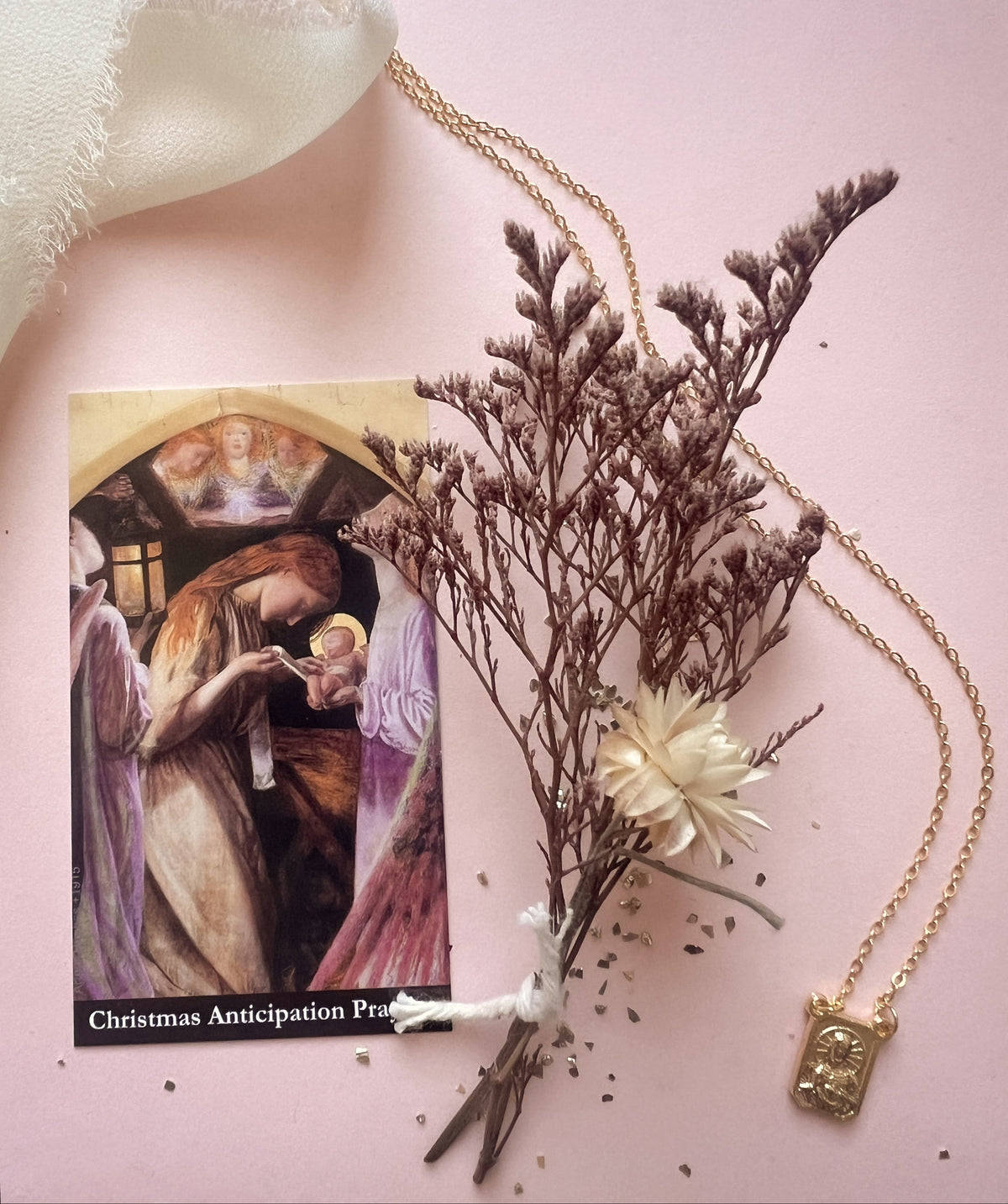 A gold-toned necklace with a small rectangular pendant is arranged on a soft pink background alongside dried brown flowers, a cream fabric corner, and a printed card titled 'Christmas Anticipation Prayer.' No person or real environment is present, so this is Product/Packaging rather than Lifestyle.