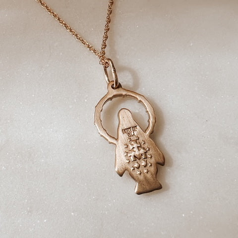 A close-up of a rose gold pendant necklace featuring a stylized fish charm with a textured body and a large circular ring detail, attached to a matching chain on a light, textured background.