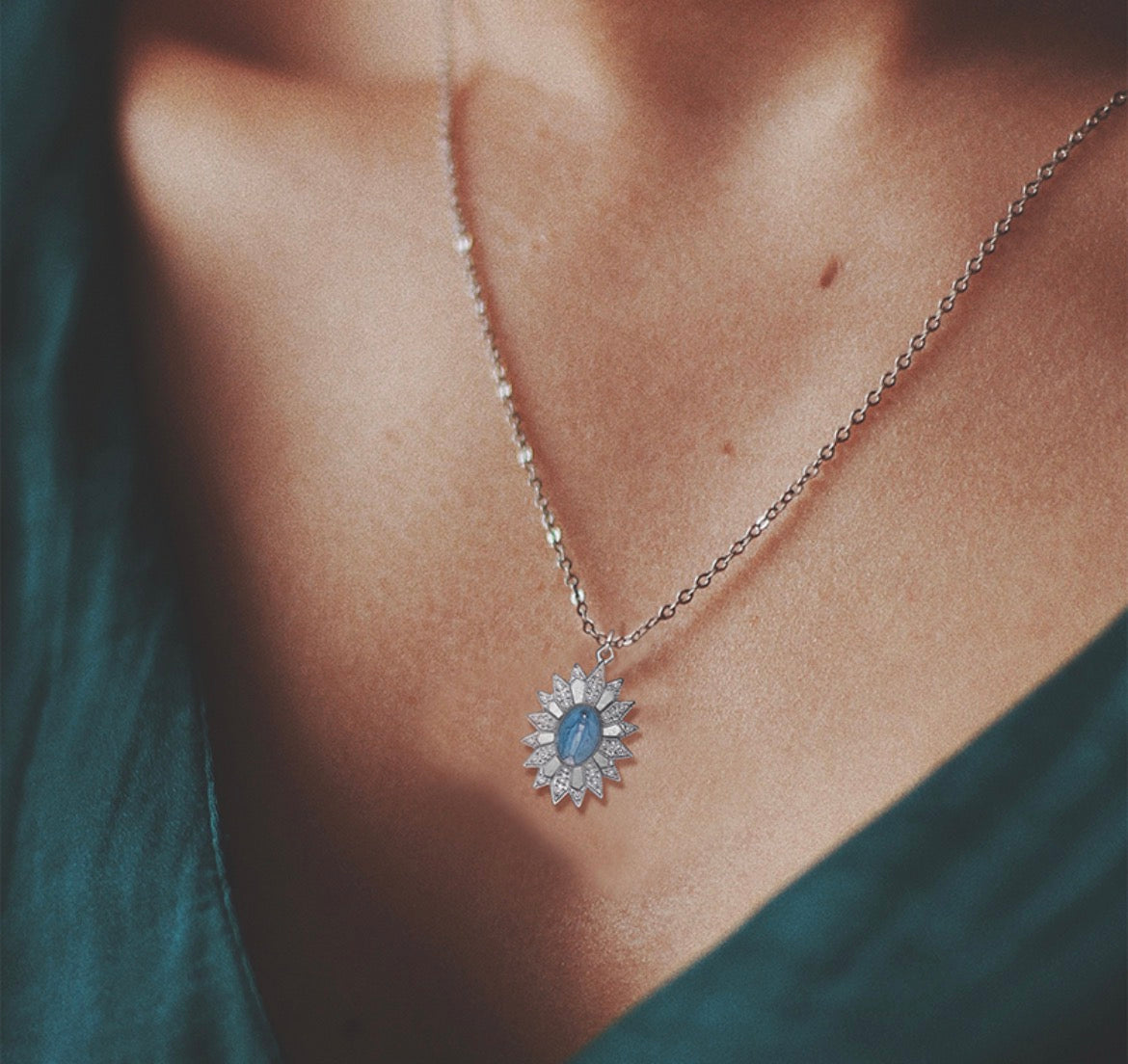 A close-up lifestyle shot of a delicate silver chain necklace with a sunburst pendant featuring a central light blue oval stone, worn by a model in a dark teal top.