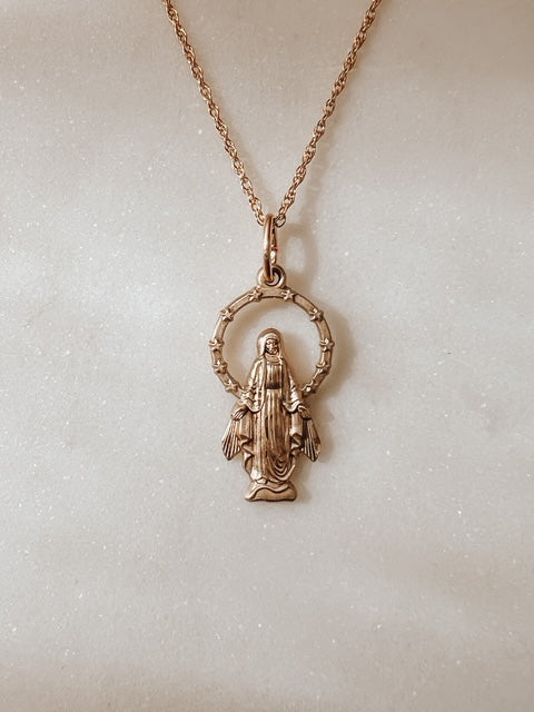 A 14k gold miraculous medal necklace featuring a small religious pendant of the Virgin Mary inside a circular halo, displayed on a textured off-white background.