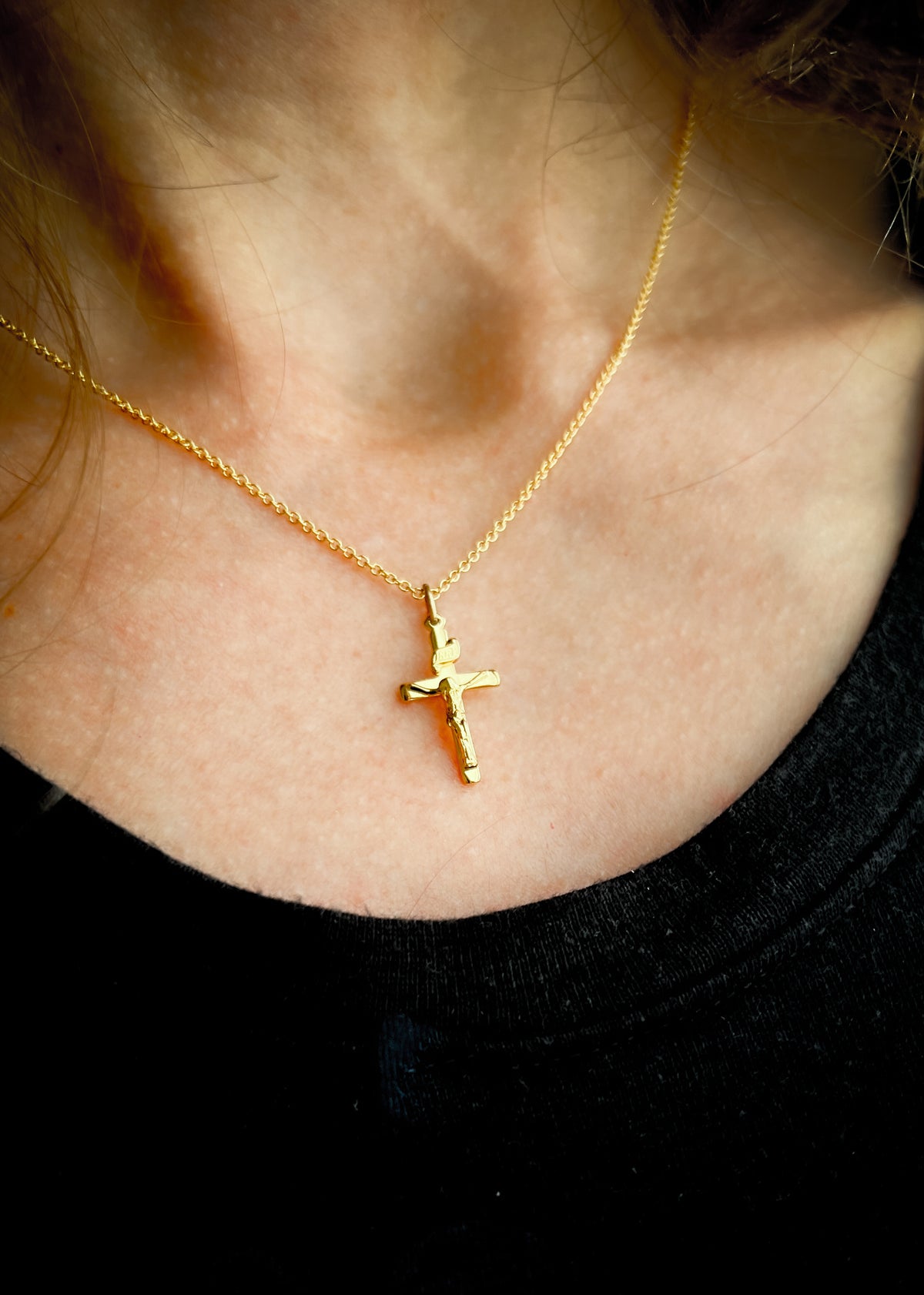 A close-up of a person's neck and chest wearing a delicate gold chain necklace with a small cross pendant, paired with a black top.