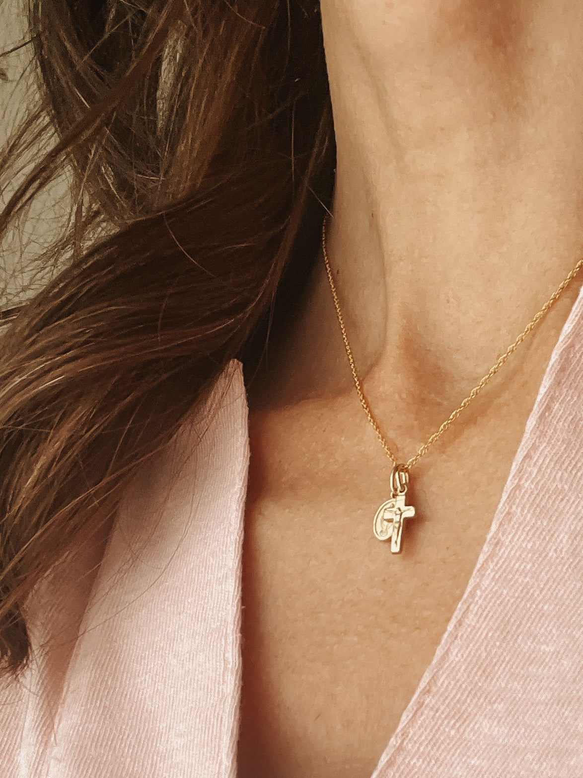 A close-up of a model wearing a delicate gold chain necklace with a small cross pendant, styled with a light pink top.