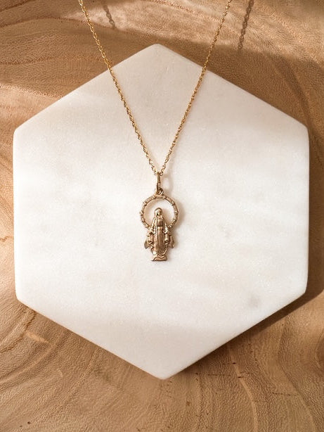 A delicate gold Miraculous medal necklace resting on a white hexagonal marble tray, set against a warm, textured wood background.